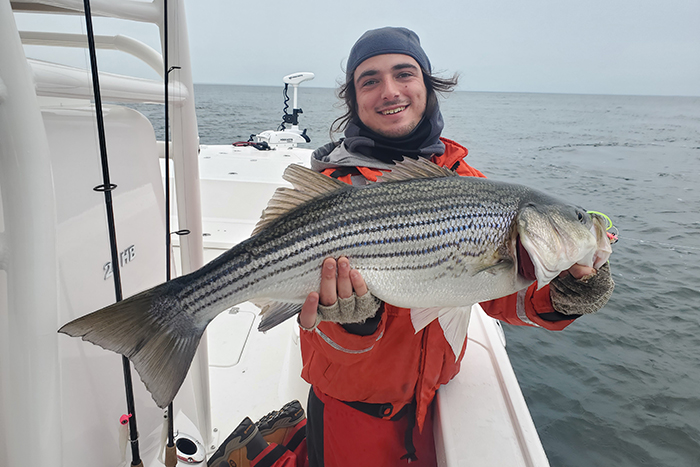 Young adult Caucasian male wearing an orange jacket and gray knit hat holding a large striped bass on a boat on the open waters