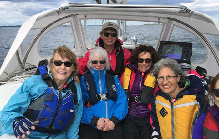 Group of 5 women all wearing bright colored jackets and with life jackets smiling in the cockpit of a sailboat on the water