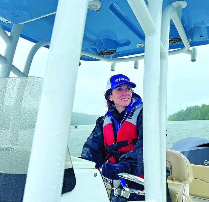 Middle-aged woman wearing a navy ballcap, navy jacket and red life jacket driving a vessel on the open waters.