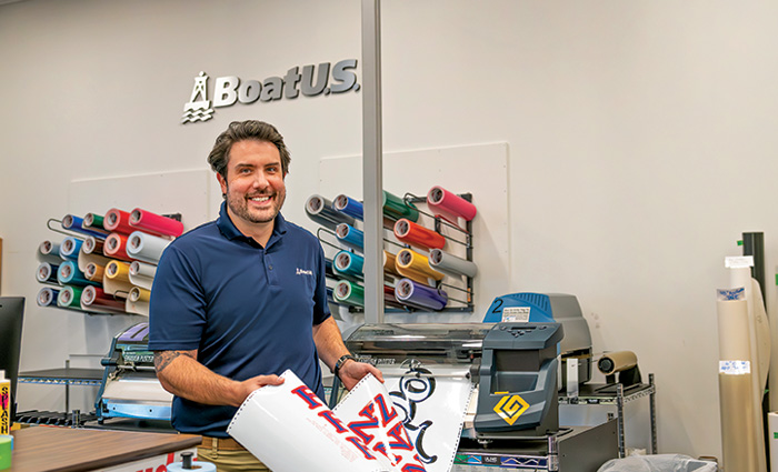 Adult male with brown hair and beard wearing a navy polo and holding a graphic in a print shop.