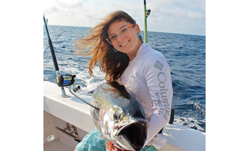 Young adult female wearing glasses and a long sleeve Columbia shirt proudly displaying a fish that was caught on a boat in open waters.
