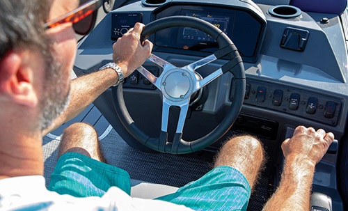 Ariel view of a an adult male wearing sunglasses and blue shorts at the wheel of a boat.