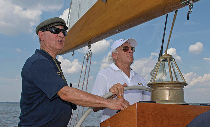 Two middle-aged men at the helm of the U.S. Naval Academy training sailboat in open waters.