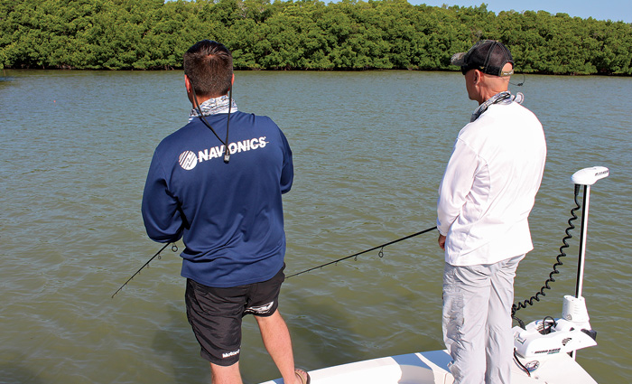A man in a navy shirt and black shorts next to a man in a white shirt and gray pants fishing off a boat.