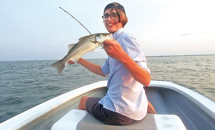 Young adult with a backwards ballcap, black rimmed glasses, gray shorts and white shirt at the front of a boat holding a fish.