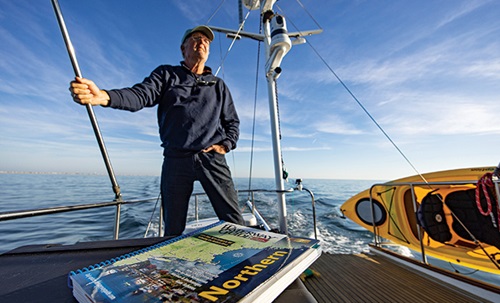 Senior adult male wearing a tan hat, dark blue jacket and pants standing at the front of a vessel on open waters.
