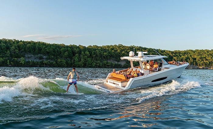 More than a  dozen passengers aboard a White Regal 38 Surf with an adult male wakeboarding on a lake. 