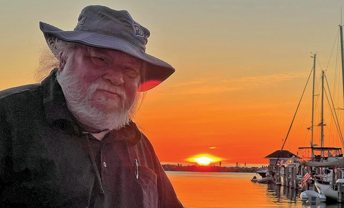 Elderly male with a gray beard, tan bucket hat and dark jacket posing for a photo in front of a sunset off the water.