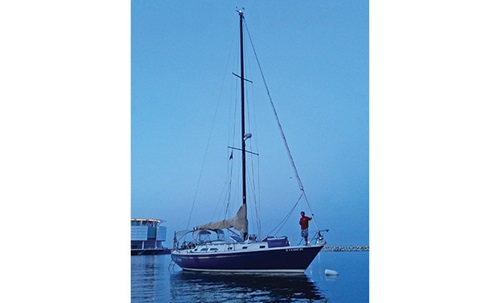 Man standing at the front of a navy and white sail boat out on the water at dusk.