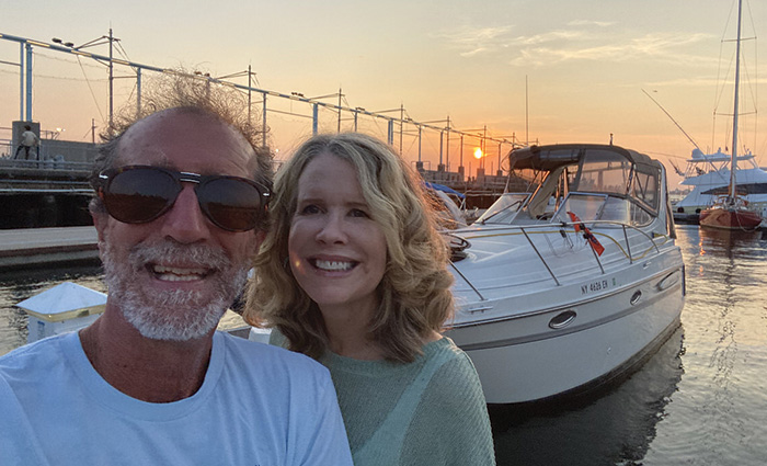 Middle-aged man with a beard wearing a white shirt and sunglasses posing with blonde female at a dock at sunset.