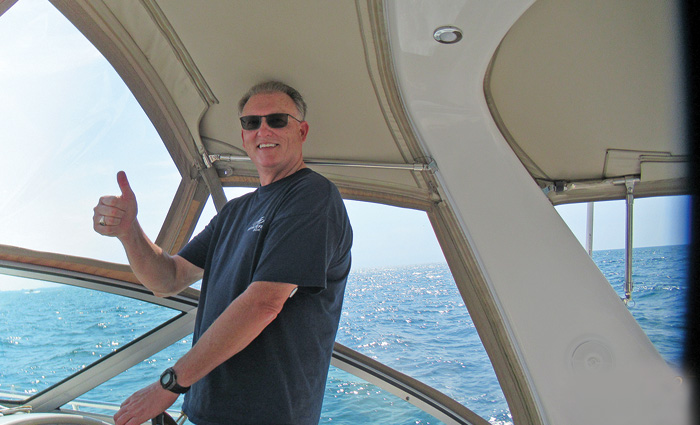 Middle-aged man wearing a navy t-shirt and sunglasses giving a 'thumbs up' while steering a boat.