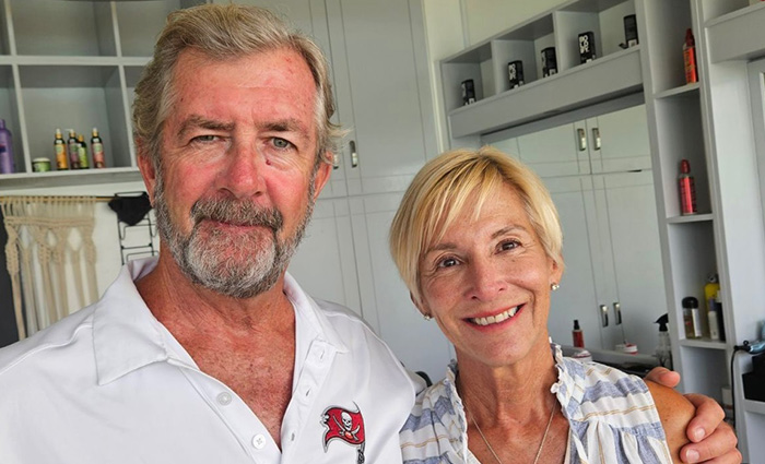 Adult male with white beard wearing a white polo posting for a picture with a blonde adult female in a kitchen.