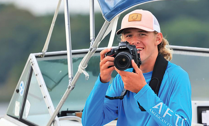 Young adult male in a blue long sleeve shirt and white hat holding a black camera.