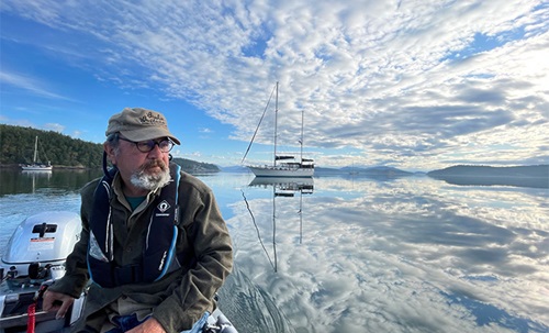 Elderly man with a white beard and tan hat navigating a small vessel on open waters where large clouds reflect off the water.