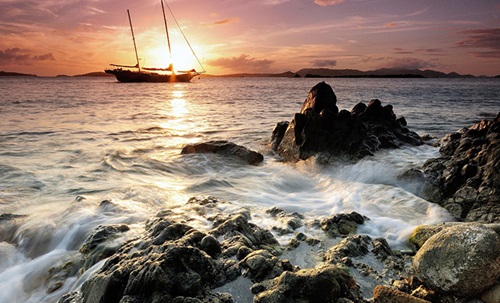 A large sailboat out on the water during sunset as waves crash upon rocks on the shore.