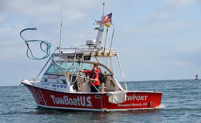 a red TowBoatUS labeled powerboat with Newport Beach, CA labels. Two men aboard boat in red shirts, one man is tossing a blue rope with seveal knots in it away from the boat